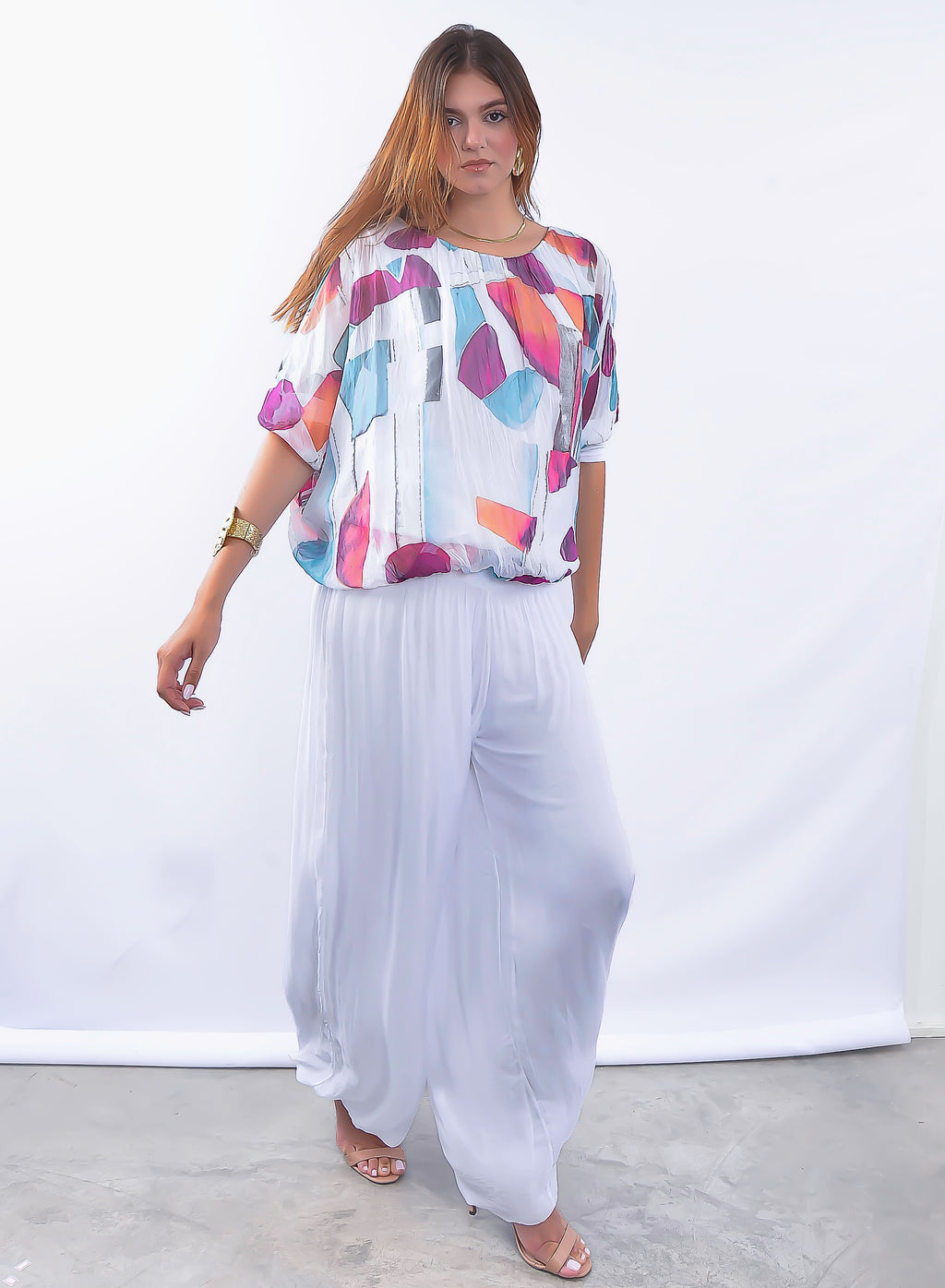 Mulher vestindo conjunto com blusa estampada colorida e calça plissada branca elegante.