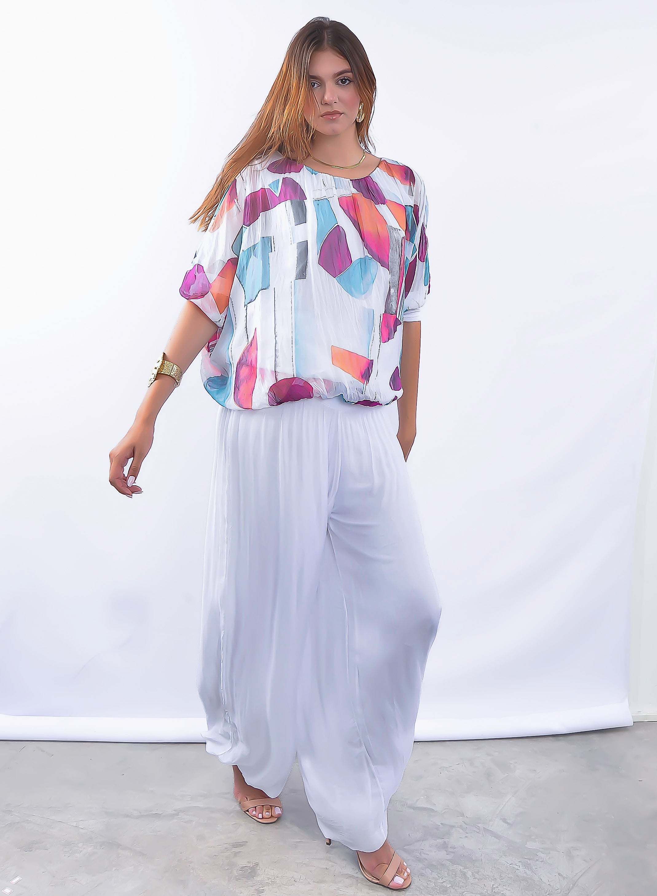 Mulher vestindo conjunto com blusa estampada colorida e calça plissada branca elegante.