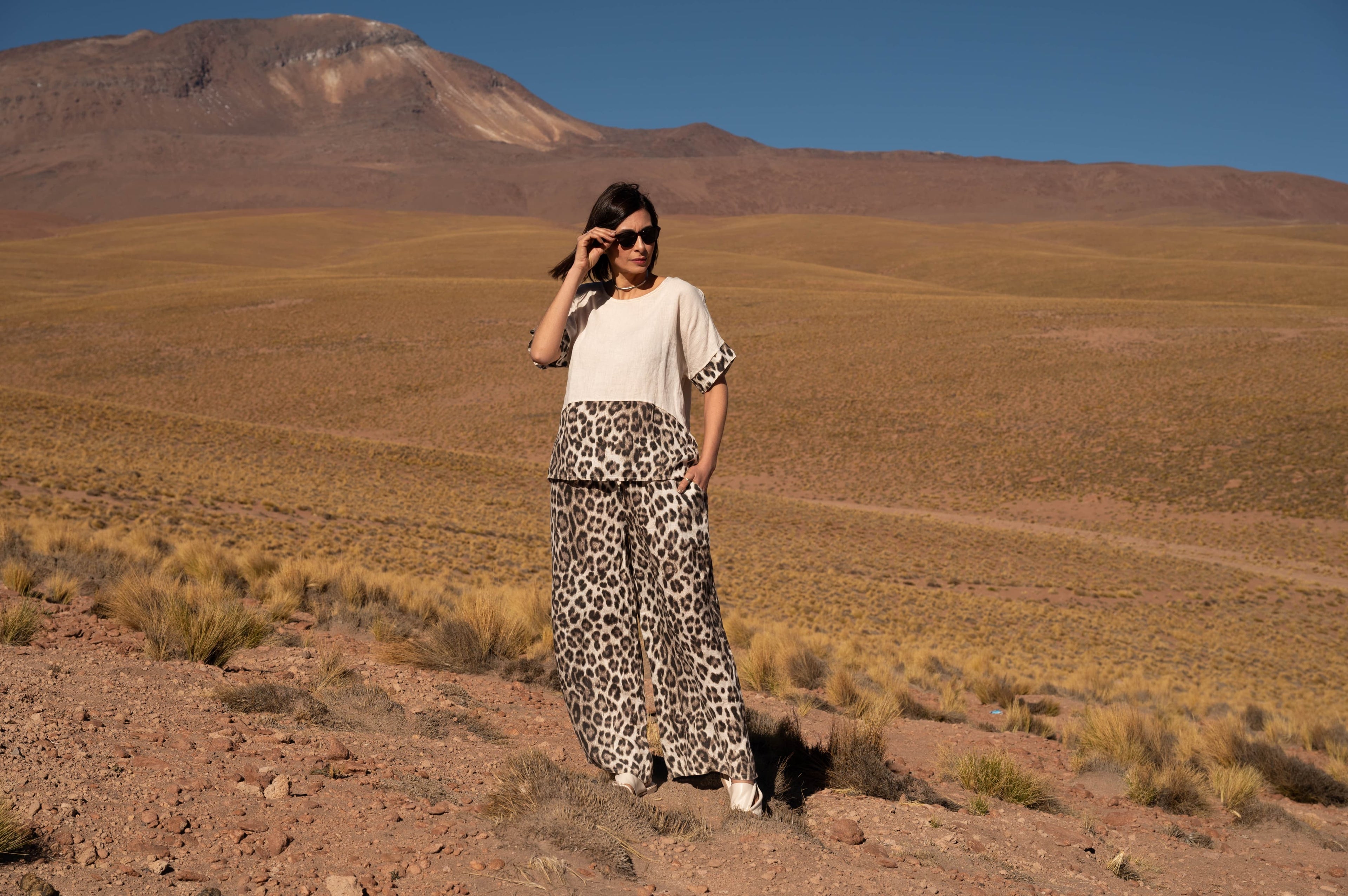 Mulher usando blusa branca e saia estampada em meio a um deserto com montanhas ao fundo