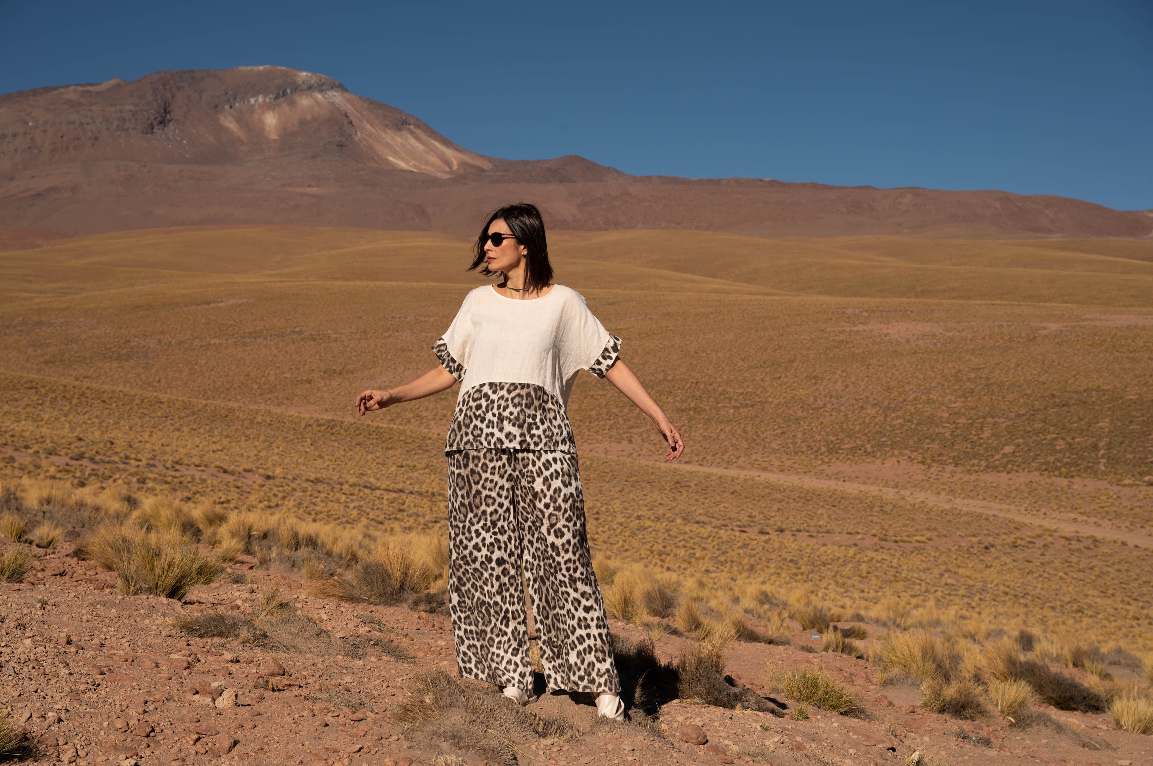 Mulher de frente no deserto, olhando para o horizonte, com roupa clara e saia longa estampada.