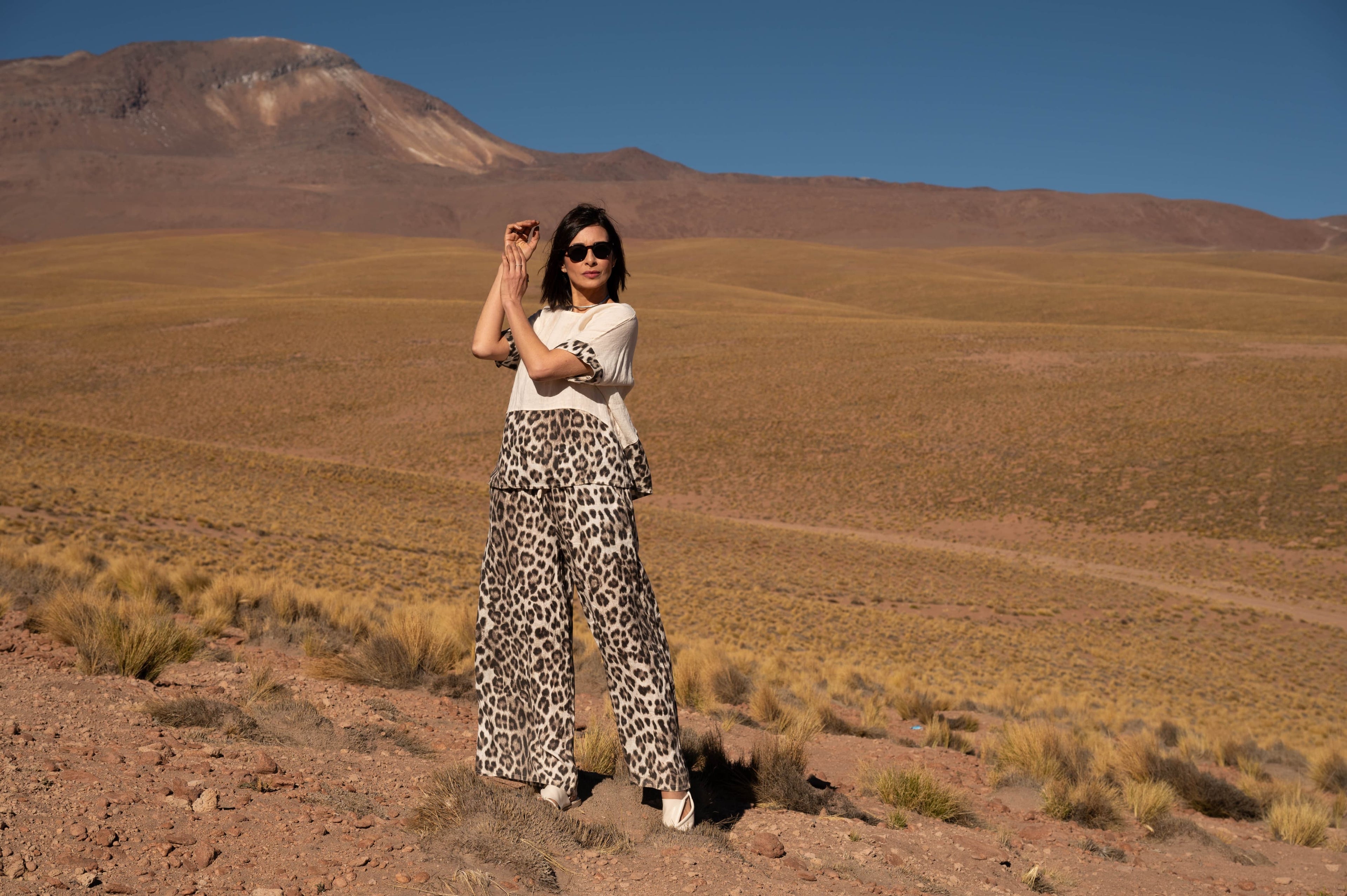 Mulher posando no deserto, com braços relaxados, vestindo blusa branca e saia longa estampada, montanhas ao fundo