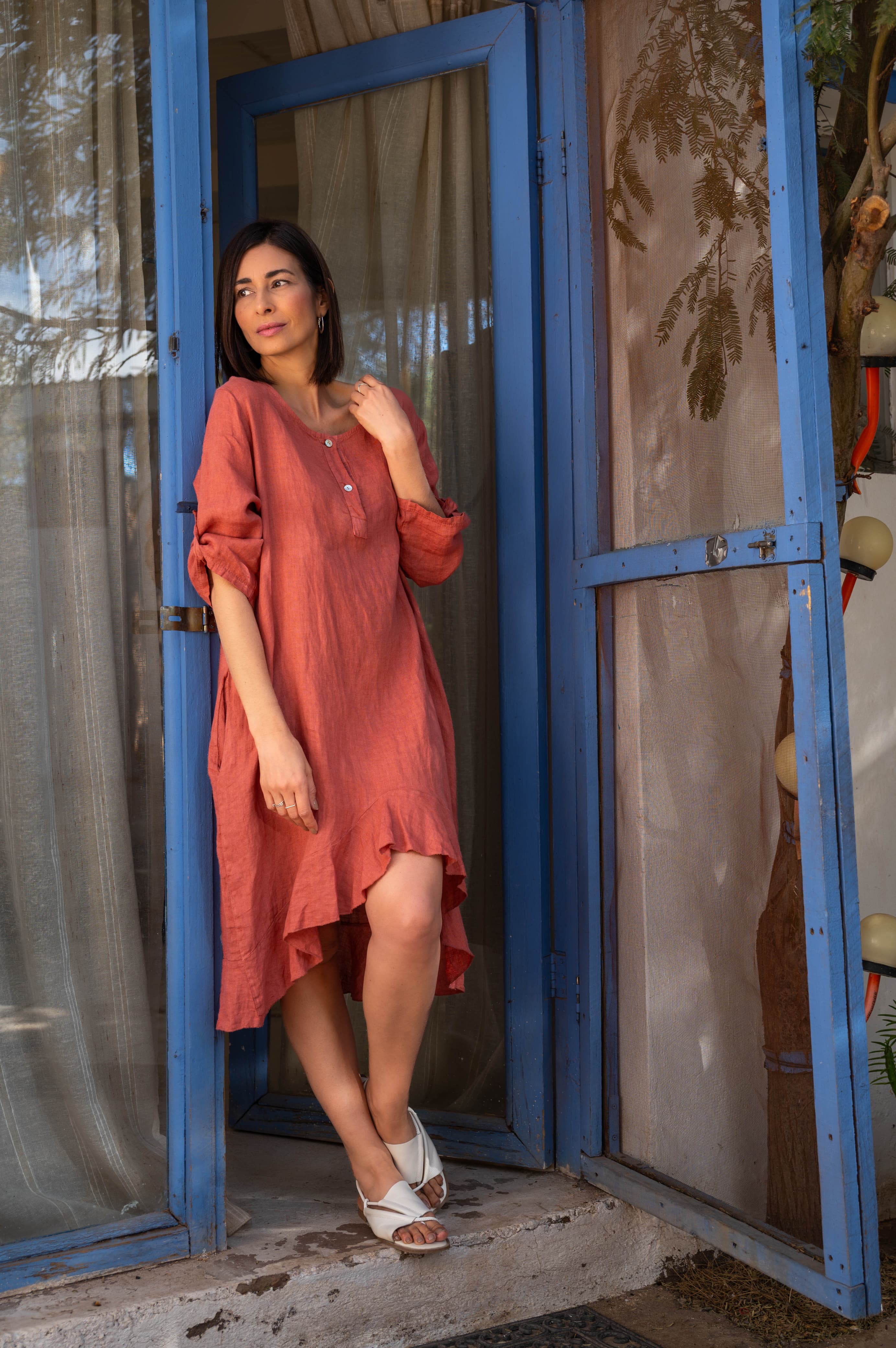 Mulher de vestido de linho terracota posando em frente a uma porta azul rústica.